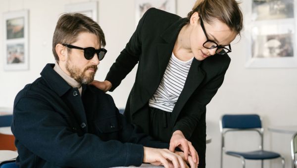 A blind man reading braille a book is aided by a standing woman in an office or classroom setting.